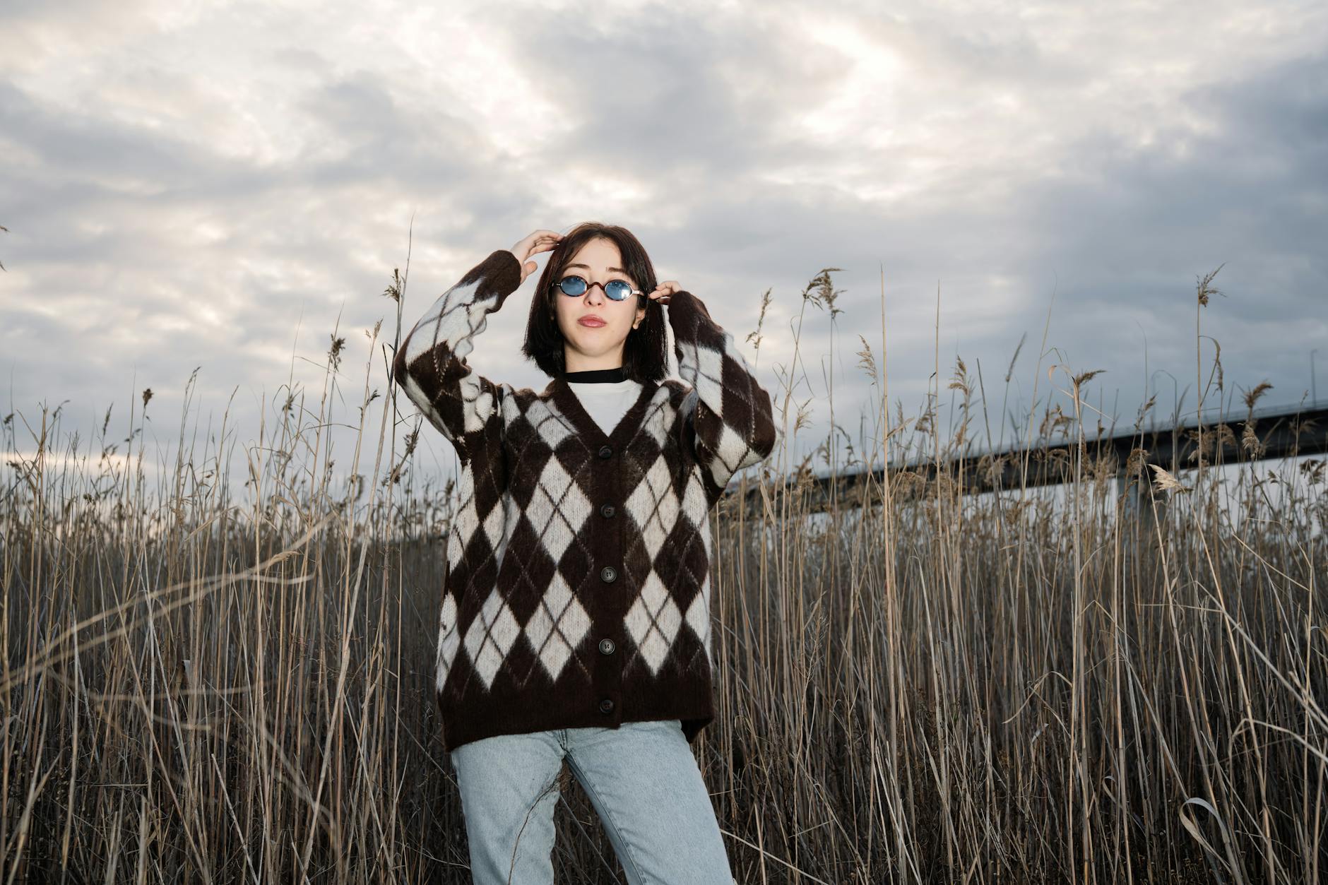 Jak nosić oversize w codziennych stylizacjach, żeby wyglądać stylowo i kobieco Kobieta w oversize swetrze argyle i okularach przeciwsłonecznych wśród traw